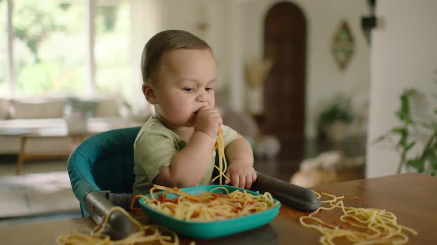 Cute baby boy sitting at a table in a booster seat eats spaghetti with his hands, making a delightful mess