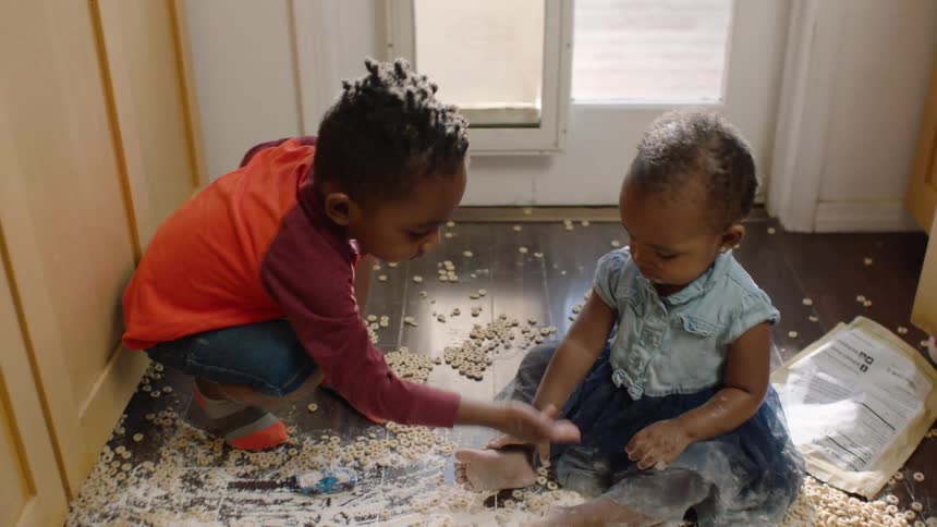 A slow-motion reveal shows two mischievous young siblings happily playing in a huge mess of spilled cereal and flour on the kitchen floor