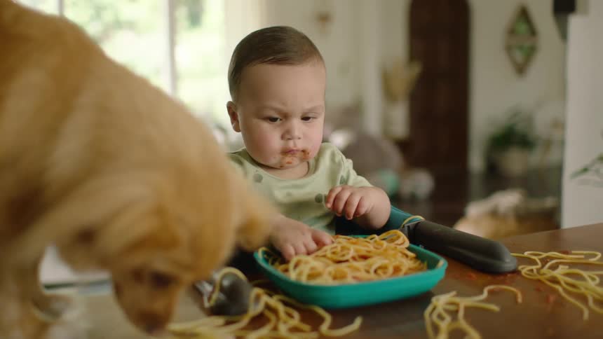 A cute baby boy eats spaghetti and makes a mess while his dog eagerly eats the dropped noodles in slow motion