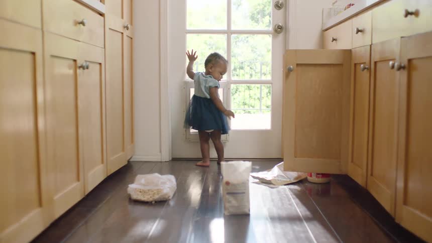A mischievous toddler looks out a sunlit kitchen door before turning away from the mess she made on the floor