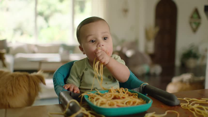 An adorable but messy baby eats spaghetti with their hands in a high chair while their hopeful dog watches