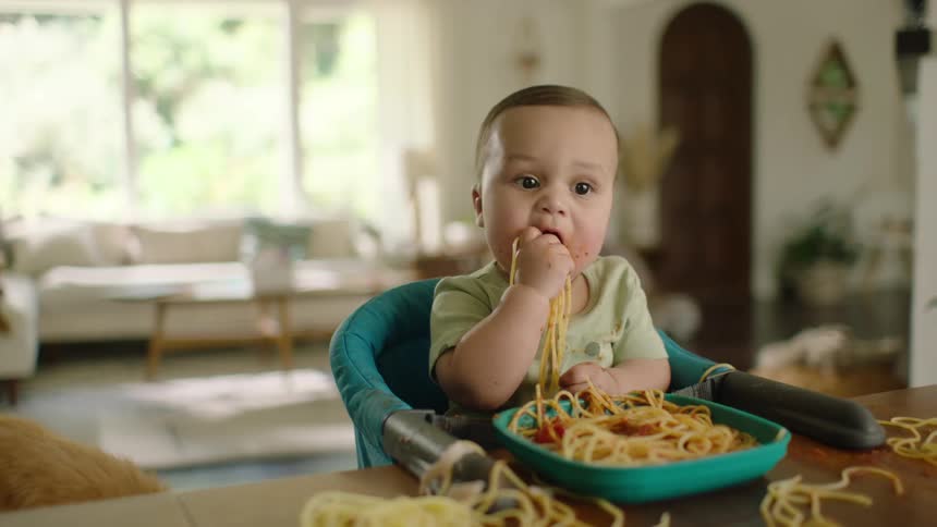 A cute and messy baby eats spaghetti with their hands while sitting in a high chair as a little dog watches and eats from the table