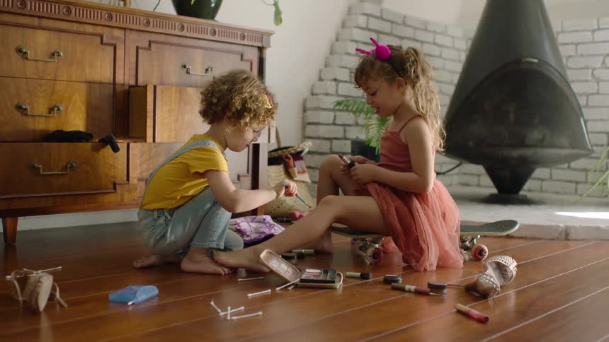A young girl sits on a skateboard applying lipstick while her younger brother paints her toes with eyeshadow in a sunlit living room