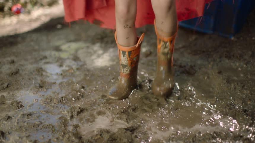 Playful young girl wearing a red cape and goggles jumps and splashes in a muddy puddle in slow motion