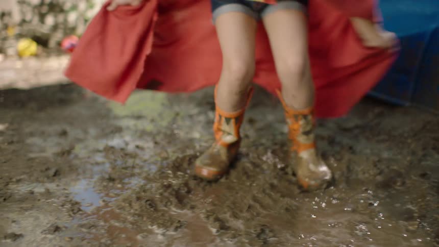 A joyful girl in a superhero costume with a red cape jumps into a muddy puddle in her backyard in slow motion