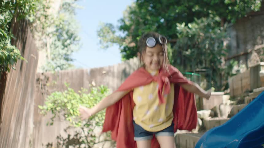 A joyful little girl wearing a red superhero cape and rain boots jumps and splashes in a muddy backyard puddle in slow motion