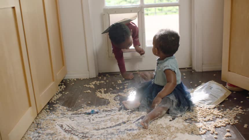 A baby girl sits in a mess of spilled cereal and flour on the kitchen floor as her older brother crawls through the pet door to play with her