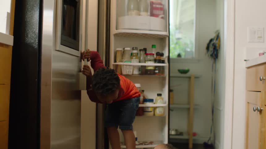 A baby girl eats blueberries on the kitchen floor while her older brother looks for a snack in the refrigerator