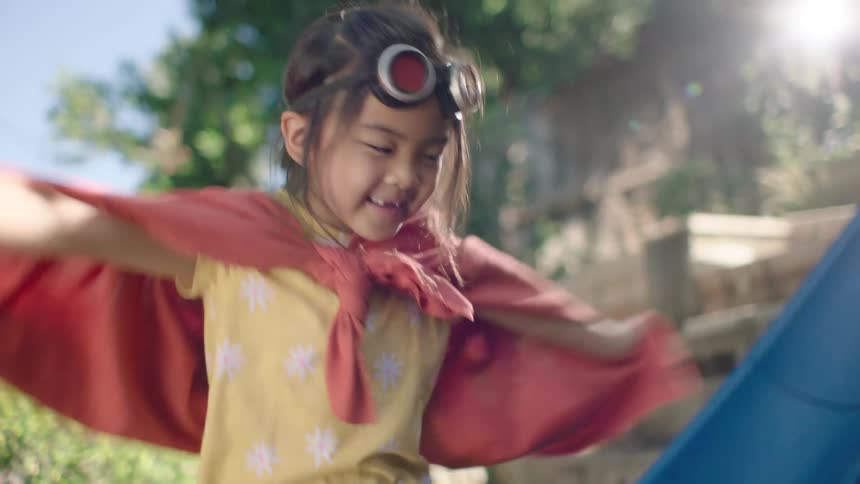 A young girl in a superhero cape and orange rain boots joyfully stomps and plays in a mud puddle in her backyard