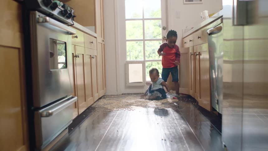 Two mischievous young siblings play together in a huge mess of spilled cereal on the kitchen floor