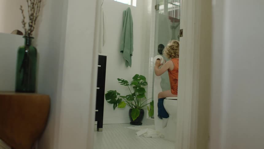 A mischievous little boy sits on the toilet and unrolls a long stream of toilet paper all over the bathroom floor