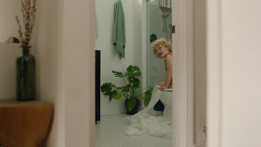 A mischievous young boy sits on the toilet and laughs as he unravels a long roll of toilet paper onto the bathroom floor