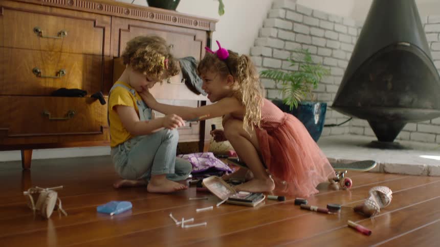 An older sister playfully applies makeup to her younger sister's face while playing dress-up on the living room floor