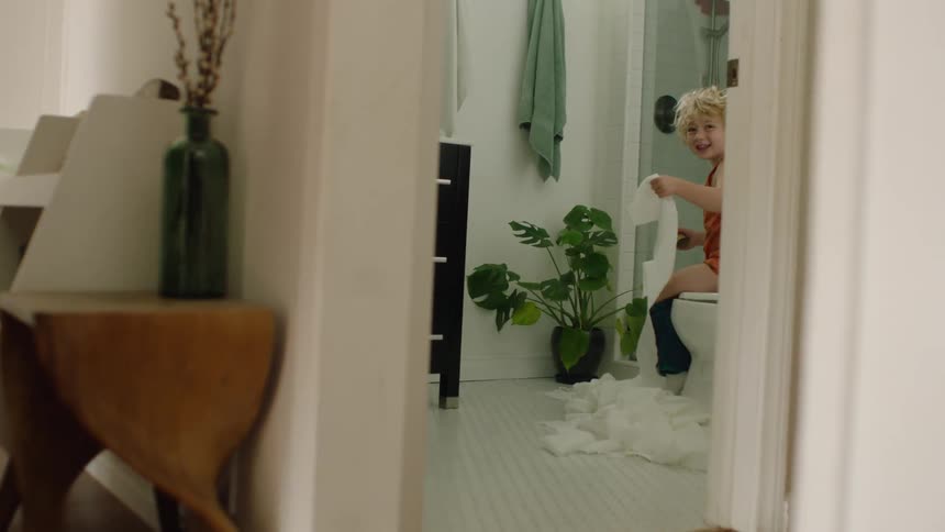 Mischievous boy sits on the toilet and happily unravels a roll of toilet paper all over the bathroom floor
