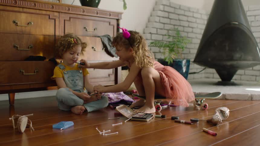 A young girl playfully applies lipstick to her sister as they play dress-up with makeup on the living room floor