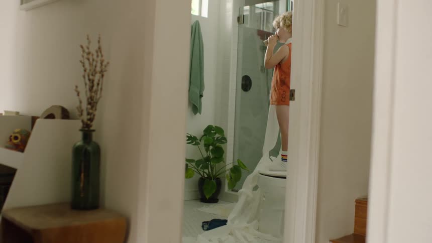 A view from the hallway shows a young boy standing on the toilet and brushing his teeth with unrolled toilet paper on the floor