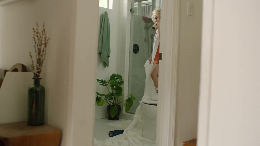 A mischievous young boy stands on the toilet, having made a mess with toilet paper, and playfully looks through the empty roll like a spyglass