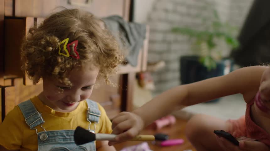 A little girl with bunny ears and a messy face playfully applies makeup to her laughing younger sister at home
