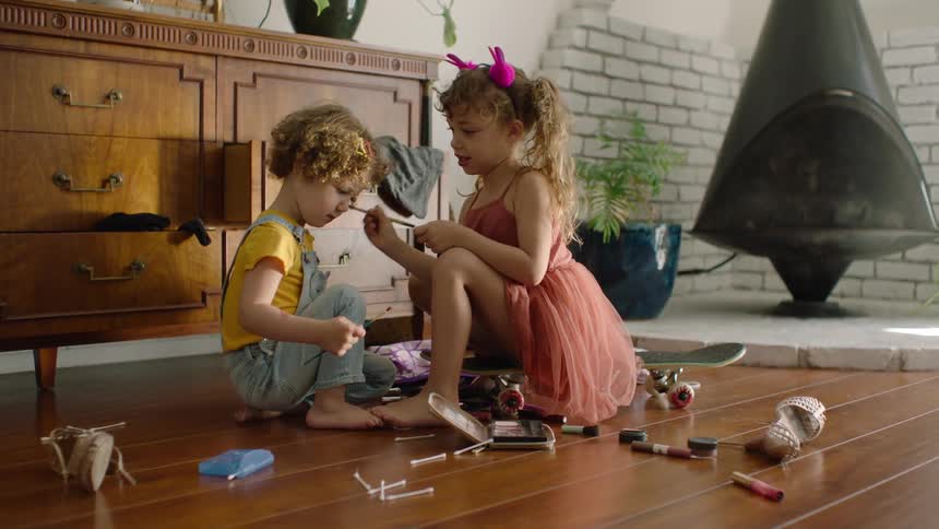 A little girl in a pink tutu sits on a skateboard and carefully applies makeup to her younger brother's face as they play on the floor