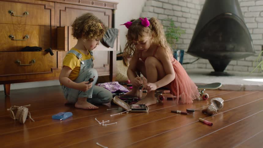A little girl in a pink dress applies makeup to her brother's face while playing dress-up on the living room floor