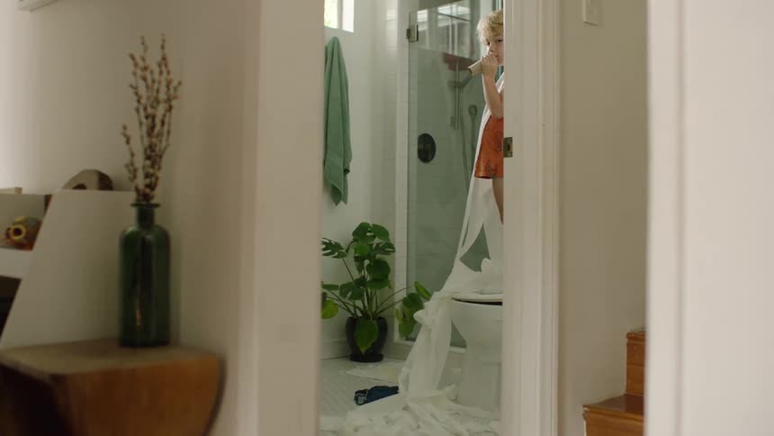 A young boy mischievously stands on the toilet and unravels the entire roll of toilet paper onto the bathroom floor