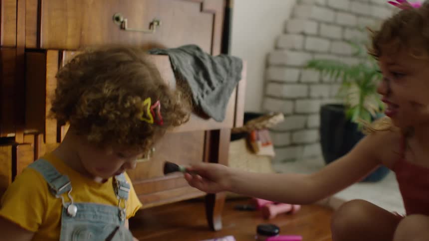 Adorable sisters with curly hair giggle while giving each other a messy makeover on the floor