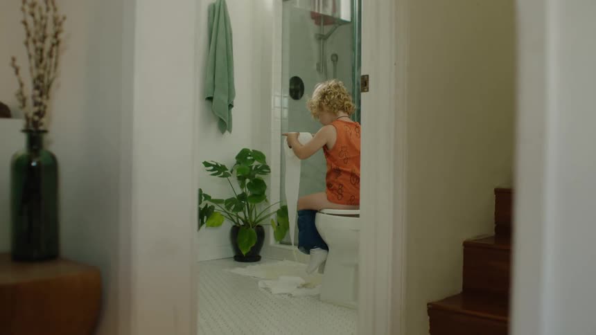 A playful little boy sits on the toilet, unrolling a long stream of toilet paper and smiling mischievously at the camera