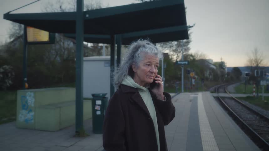 A mature woman with long gray hair talks on the phone and smiles while waiting at a train station