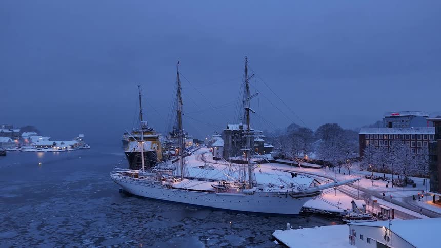 A majestic snow-covered tall ship is docked in an icy Scandinavian harbor during a cold winter evening