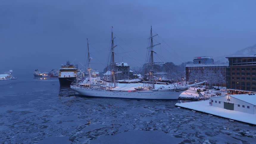 Aerial view of a historic sailing ship covered in snow, docked in an icy harbor surrounded by city buildings at twilight