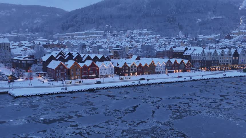 Aerial view of the historic snow-covered Bryggen wharf in Bergen, Norway, with its colorful buildings lit up beside the icy harbor at dusk