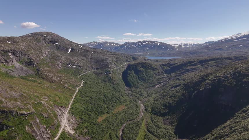 A static high-angle aerial view captures a vast mountain landscape with a winding road through a lush green valley