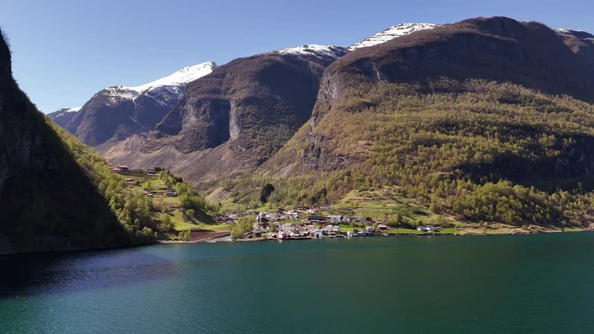 A tranquil village sits on the edge of a serene fjord in Norway, surrounded by towering mountains with snow-capped peaks under a bright blue sky
