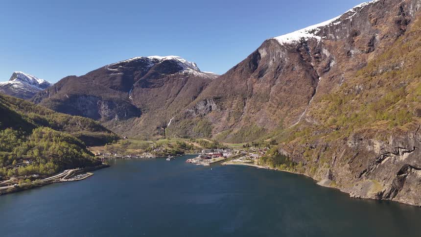Static aerial shot captures the stunning beauty of a Norwegian village at the head of a tranquil fjord, enclosed by towering, snow-dusted mountains under a clear blue sky