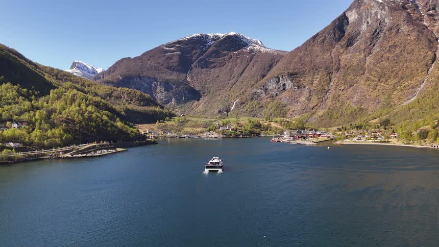 A modern passenger ferry sails through a majestic Norwegian fjord, approaching a scenic village nestled between towering, snow-capped mountains on a sunny day