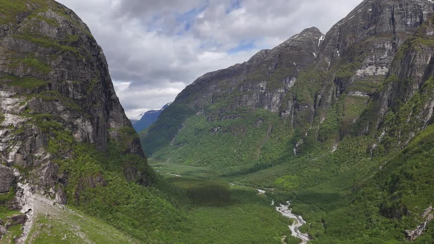 A majestic aerial pullback shot over a verdant river valley nestled between towering, dramatic mountains in the Norwegian wilderness