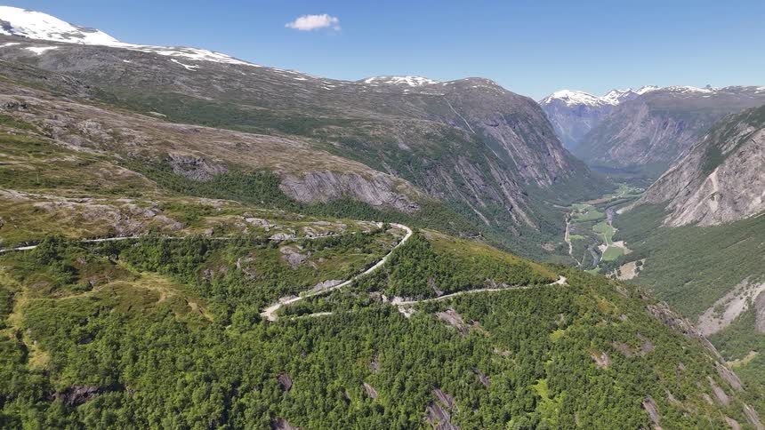 Slow aerial pullback reveals a winding road through a majestic green mountain valley with snow-capped peaks