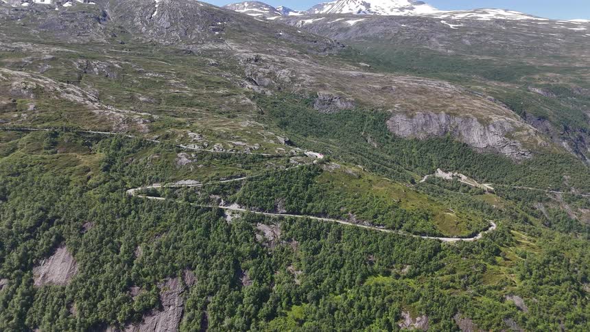 A static aerial view of a winding serpentine road snaking through a green mountain valley with snow-capped peaks in the distance