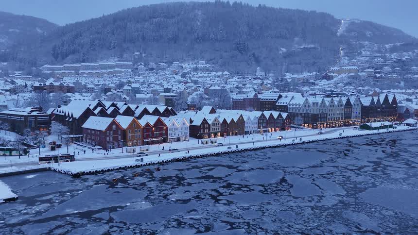 A picturesque winter view of the snow-covered historic Bryggen wharf in Bergen, Norway, with its colorful wooden houses lit up at twilight as ice floes drift in the harbor