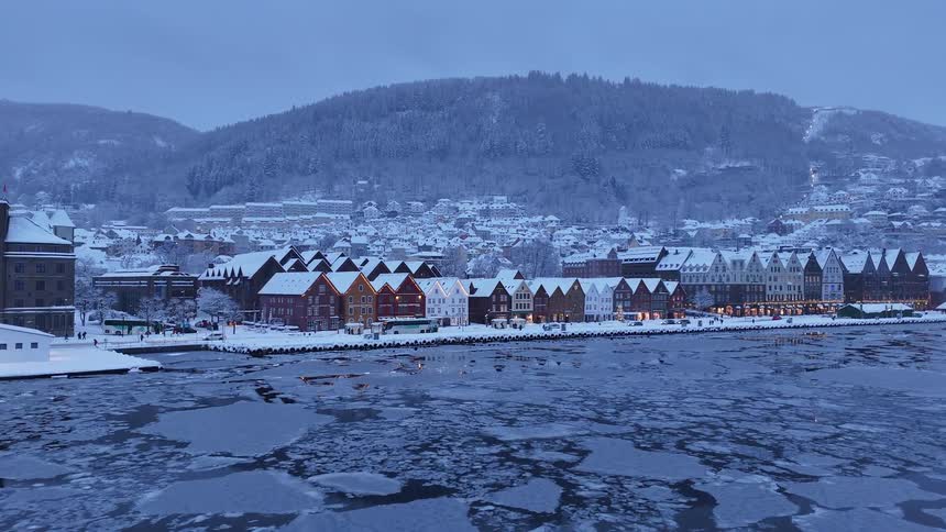 A tranquil winter scene of Bergen's historic Bryggen waterfront, with snow-covered buildings and mountains surrounding the icy harbor at dusk