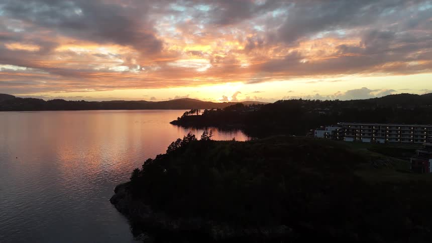 An aerial view of a tranquil fjord at sunset, with colorful clouds reflecting on the water and silhouetted hills with houses