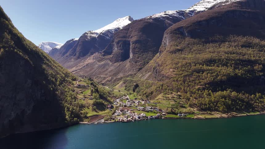 A stunning aerial view captures a small village nestled on the edge of a fjord, surrounded by majestic, snow-capped mountains in Norway