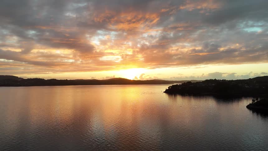 An aerial pullback reveals a glorious golden sunset casting its warm light over a tranquil body of water and a silhouetted coastline