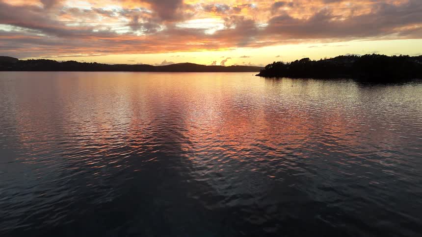 A serene aerial view tilts up from calm waters to reveal a breathtaking, fiery sunset over a vast lake