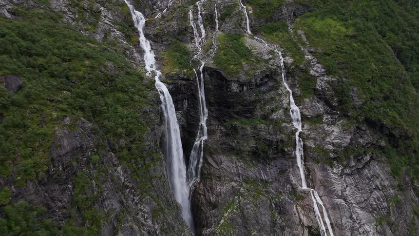 An epic aerial view zooms out from multiple waterfalls flowing down a steep, green mountainside in a remote wilderness