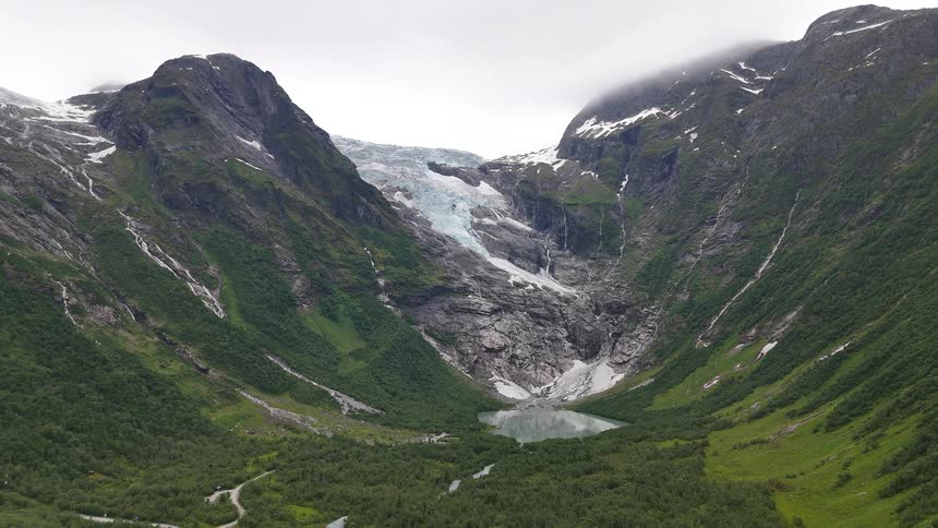 A slow aerial pullback reveals the magnificent Bondhusbreen glacier, its glacial lake, and the lush green valley below in Norway