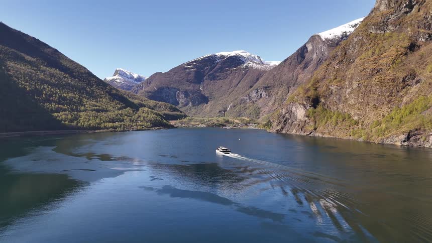 A boat sails through a magnificent Norwegian fjord, surrounded by steep mountains with snow-capped peaks under a clear blue sky