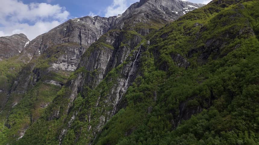 A stunning view of a waterfall on a verdant mountainside, zooming out to show the towering, snow-dusted peaks