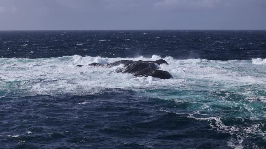 Slow motion footage shows powerful, foamy ocean waves crashing over a large rock in the deep blue sea