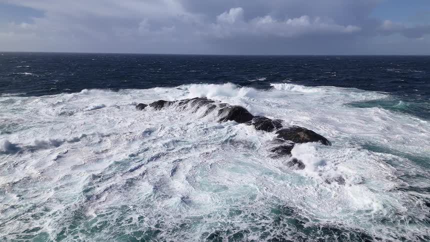 A dramatic slow-motion aerial shot of powerful ocean waves crashing over dark rocks in a turbulent sea under a cloudy sky